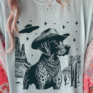 May include: A light gray t-shirt featuring a black and white illustration of a dog wearing a cowboy hat and bandana. The design includes a desert landscape with cacti, rock formations, and a UFO. The dog has a spotted coat.