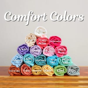 May include: A pyramid of rolled-up fabric in various colors, including white, ivory, violet, and terracotta. The image features the text "Comfort Colors" at the top, with each roll labeled with its color name. The fabric rolls are neatly stacked.