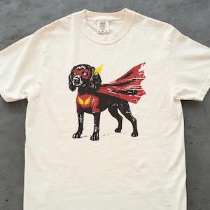 May include: Cream-colored t-shirt featuring a graphic of a black dog dressed as a superhero. The dog wears a red mask, cape, and chest emblem with yellow wings. The shirt has a crew neck and short sleeves.