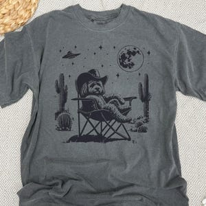 Op de afbeelding: Een donkergrijs T-shirt met een grafische print van een hond die een cowboyhoed draagt en ontspant in een campingstoel onder een halve maan en sterren. De afbeelding bevat ook cactussen en een UFO.
