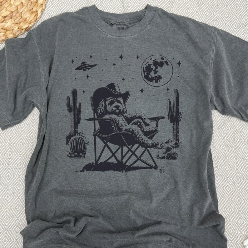 Op de afbeelding: Een donkergrijs T-shirt met een grafische print van een hond die een cowboyhoed draagt en ontspant in een campingstoel onder een halve maan en sterren. De afbeelding bevat ook cactussen en een UFO.
