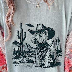 Pode incluir: Camiseta verde claro com um design gráfico em preto e branco. O design apresenta um cão com chapéu de cowboy e bandana, num cenário desértico com um cacto e um OVNI.