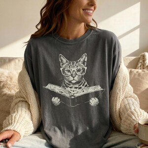 May include: A charcoal gray crew neck t-shirt featuring a white graphic print of a cat wearing glasses and reading an open book. The cat is centered on the chest, depicted with detailed line work, and the shirt has a relaxed, loose-fitting silhouette.