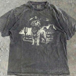 May include: A charcoal grey short-sleeve t-shirt with a vintage wash finish. The center graphic features a dog wearing a cowboy hat and bandana, standing in a desert landscape with a saguaro cactus, rock formations, and a flying saucer above.