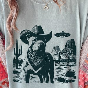 Könnte beinhalten: Hellblaues T-Shirt mit einem Motiv eines Hundes mit Cowboyhut und Halstuch, vor einer Wüstenlandschaft mit Kakteen und einem UFO. Das Design ist in Schwarz und Grau gehalten.