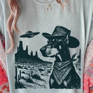 May include: A light gray t-shirt featuring a black and white graphic of a Doberman Pinscher wearing a cowboy hat and bandana, with a desert landscape and UFOs in the background. The design has a vintage, western theme.