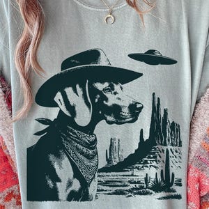 May include: A light gray t-shirt featuring a black and white graphic of a dog wearing a cowboy hat and bandana, with a desert landscape and a UFO in the background. The dog is a Weimaraner breed.