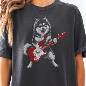 Pode incluir: Uma t-shirt de manga curta cinzento-carvão com uma estampa gráfica de um cão fofo de pé nas patas traseiras a tocar uma guitarra elétrica vermelha. O cão tem pelo preto e branco e a guitarra tem uma placa de proteção branca.