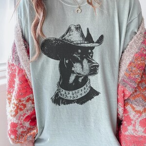 Pu&ograve; includere: T-shirt azzurra con grafica in bianco e nero di un cane con cappello da cowboy e collare borchiato. Il ritratto del cane &egrave; centrato sulla maglietta, creando un design audace e accattivante.