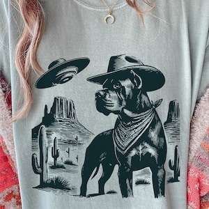 May include: A light gray t-shirt featuring a black graphic of a dog wearing a cowboy hat and bandana, with a UFO above. The dog is set against a desert landscape with cacti and mountains. The shirt is a casual wear item.