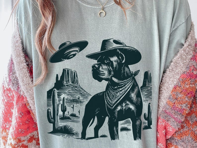 May include: A light gray t-shirt featuring a black graphic of a dog wearing a cowboy hat and bandana, with a UFO above. The dog is set against a desert landscape with cacti and mountains. The shirt is a casual wear item.