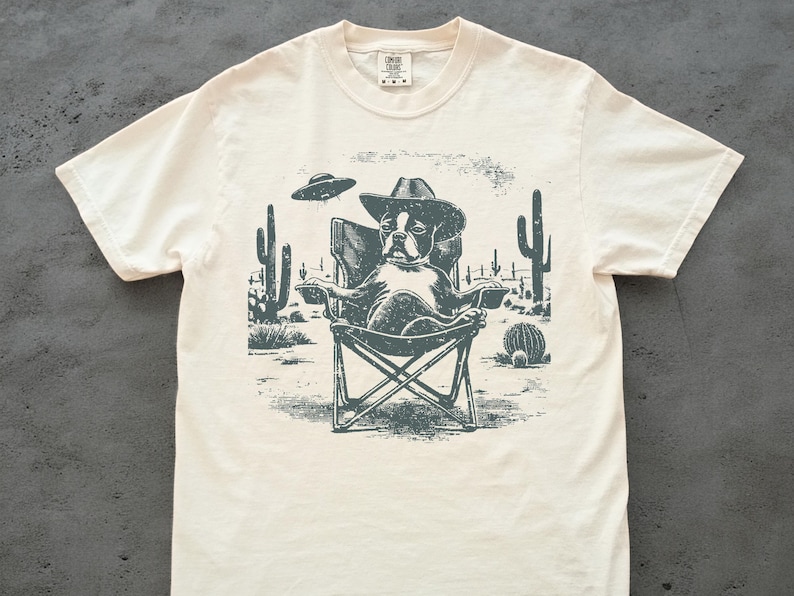 May include: Cream-colored t-shirt featuring a graphic of a dog wearing a cowboy hat, seated in a folding chair in a desert scene with cacti and a UFO. The design is in shades of gray and green.