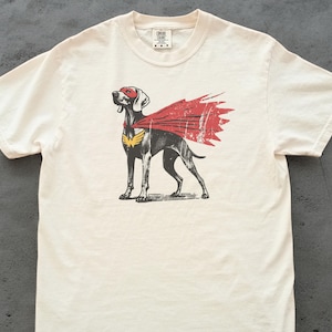 Puede incluir: Camiseta color crema con un diseño de un perro con disfraz de superhéroe. El perro tiene una máscara y capa rojas, con un emblema amarillo en el pecho. El diseño tiene un aspecto vintage.