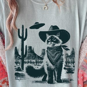 Puede incluir: Camiseta gris claro con un diseño gráfico en blanco y negro de un gato con sombrero de vaquero y pañuelo, sobre un paisaje desértico con un OVNI. El gato es el elemento central del diseño.