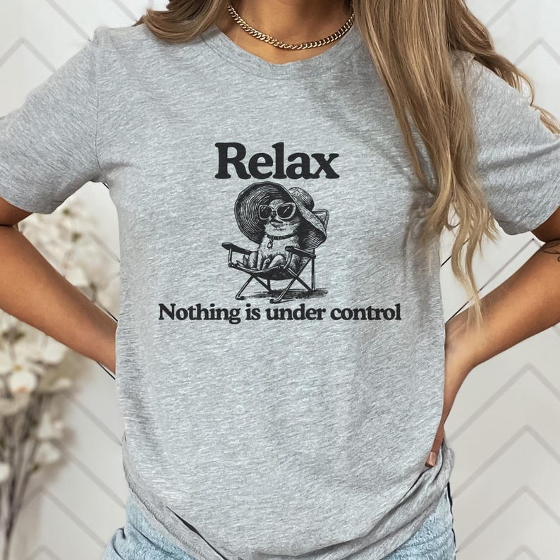 May include: A heather gray t-shirt featuring the word "Relax" above an illustration of a cat wearing sunglasses and a sun hat, seated in a beach chair. The text "Nothing is under control" is below the illustration.