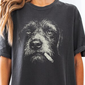 May include: A charcoal gray short-sleeve t-shirt featuring a large, detailed graphic print of a dog's face in a light cream color. The dog is depicted with a cigarette in its mouth, rendered in a high-contrast, sketch-like artistic style.