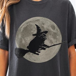 Könnte beinhalten: Dunkelgraues T-Shirt mit einem schwarzen Mops, der einen Hexenhut trägt und auf einem Besen vor einem Vollmond reitet. Ein Halloween-Design, ideal für Hundeliebhaber.