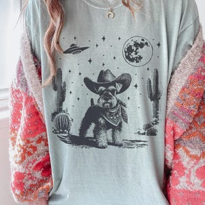 May include: A light blue t-shirt featuring a black and white illustration of a dog wearing a cowboy hat and bandana, set against a desert landscape with cacti, a full moon, and a UFO. The shirt has a relaxed fit.