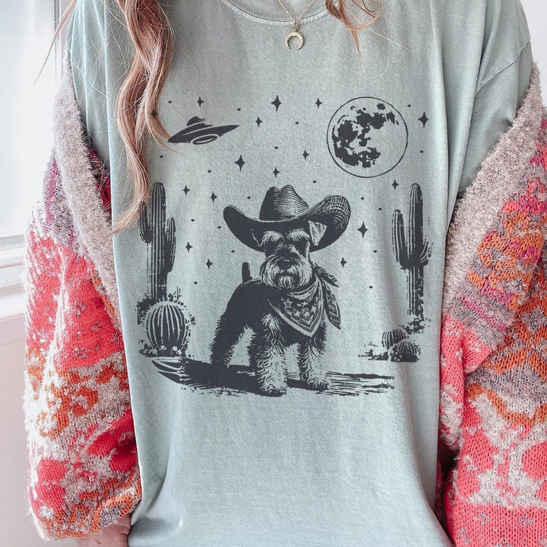 May include: A light blue t-shirt featuring a black and white illustration of a dog wearing a cowboy hat and bandana, set against a desert landscape with cacti, a full moon, and a UFO. The shirt has a relaxed fit.
