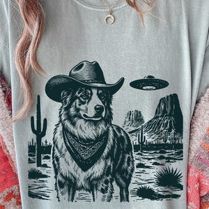 May include: A light gray t-shirt featuring a black and white illustration of a dog wearing a cowboy hat and bandana, with a desert landscape and a UFO in the background. The design includes cacti and mountain silhouettes.