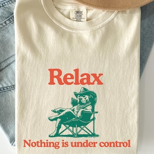 Pode incluir: Camiseta creme com a palavra "Relax" em letras laranja. Abaixo, uma ilustração verde de um cachorro usando um chapéu de cowboy e sentado em uma cadeira dobrável. O texto "Nothing is under control" está abaixo da ilustração.