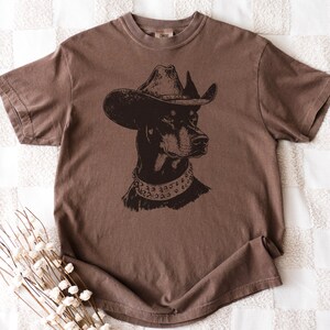 Pu&ograve; includere: T-shirt marrone con grafica nera di un Dobermann Pinscher con cappello da cowboy e collare borchiato. La maglietta ha girocollo e maniche corte. Il ritratto del cane &egrave; centrato.