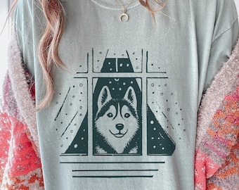 May include: A light blue t-shirt features a graphic of a Husky dog looking out a window. The dog is black and white, with a smiling expression. The window is framed with a dark green border, and the background has a starry night design.