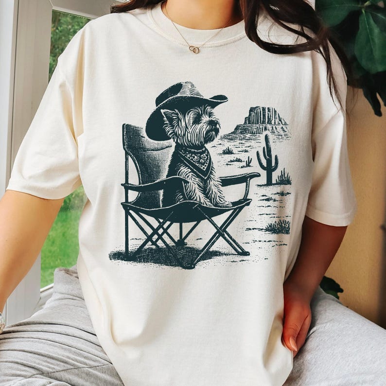 Pu&ograve; includere: T-shirt color crema con un'illustrazione in bianco e nero di un cane con cappello da cowboy e bandana, seduto su una sedia pieghevole. Lo sfondo raffigura un paesaggio desertico con una montagna e un cactus.