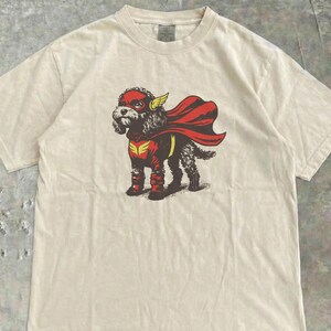 Puede incluir: Camiseta color crema con un diseño de un perro disfrazado de superhéroe. El perro lleva una máscara, capa y botas rojas, con detalles amarillos. La camiseta tiene cuello redondo y mangas cortas. El diseño está impreso en la parte delantera.