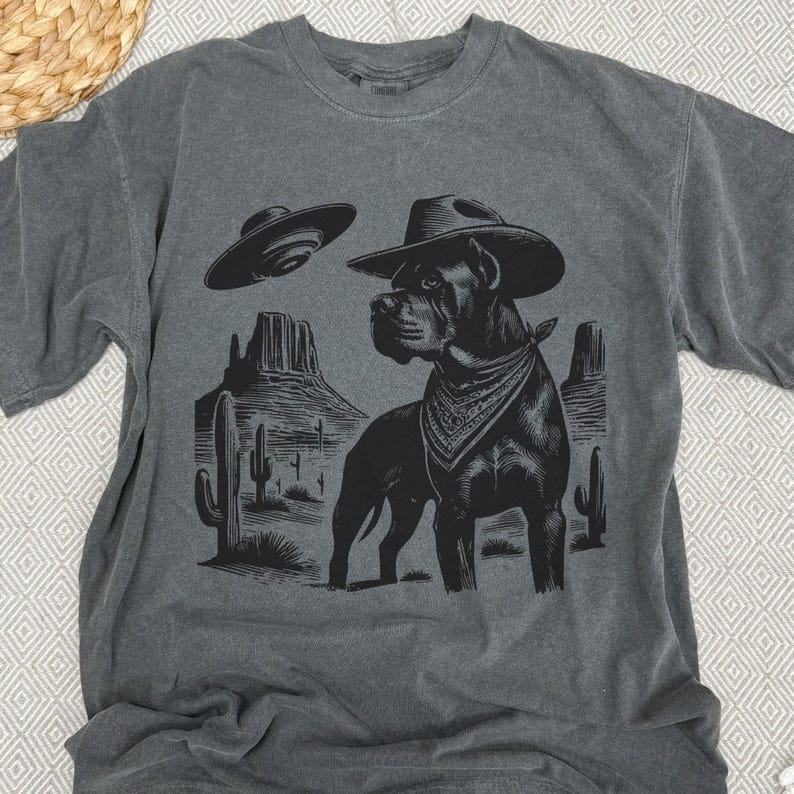 May include: Dark gray t-shirt featuring a black graphic of a dog wearing a cowboy hat and bandana, set against a desert landscape with cacti and a UFO. The design has a vintage, distressed look.