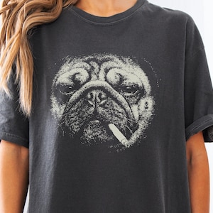 May include: A charcoal gray short-sleeve t-shirt featuring a large, detailed white graphic print of a pug dog face. The pug is depicted with a cigarette in its mouth, rendered in a high-contrast, stippled artistic style centered on the chest.