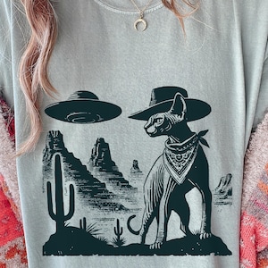 Pode incluir: Camiseta azul clara com um desenho em verde escuro. O desenho mostra um gato Sphynx com chapéu de cowboy e bandana, com um OVNI sobre uma paisagem desértica.