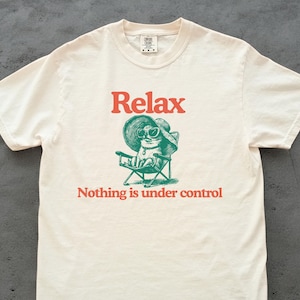 May include: Cream-colored t-shirt with the word "Relax" in red above a green illustration of a cat wearing sunglasses and a hat, seated in a folding chair. Below the illustration, the text reads "Nothing is under control."