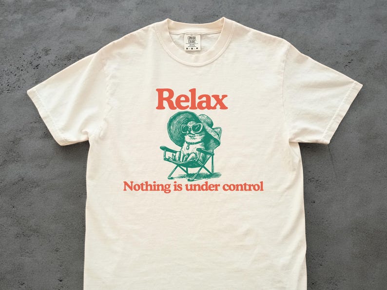 May include: Cream-colored t-shirt with the word "Relax" in red above a green illustration of a cat wearing sunglasses and a hat, seated in a folding chair. Below the illustration, the text reads "Nothing is under control."