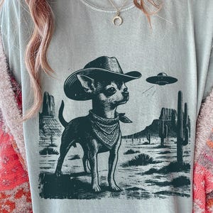May include: A light gray t-shirt featuring a black and white illustration of a Chihuahua wearing a cowboy hat and bandana, with a UFO in the background. The design includes desert scenery.