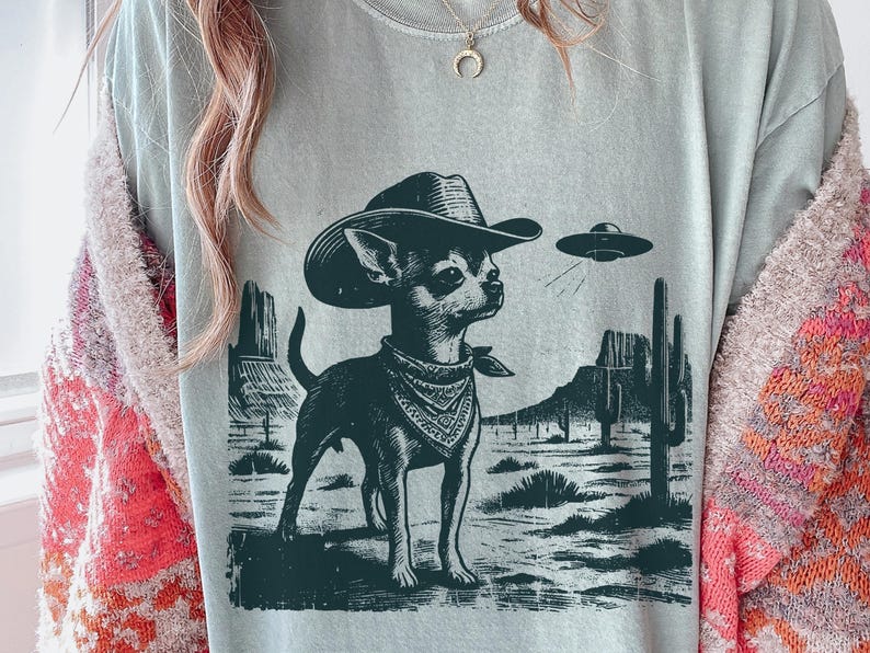May include: A light gray t-shirt featuring a black and white illustration of a Chihuahua wearing a cowboy hat and bandana, with a UFO in the background. The design includes desert scenery.