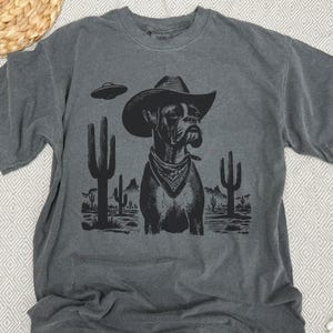 Puede incluir: Camiseta gris oscuro con un dise&ntilde;o negro de un perro Boxer con sombrero de vaquero y pa&ntilde;uelo, con un OVNI y un paisaje des&eacute;rtico. El dise&ntilde;o incluye cactus. La camiseta tiene cuello redondo y mangas cortas.