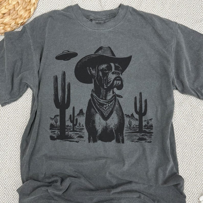 Puede incluir: Camiseta gris oscuro con un dise&ntilde;o negro de un perro Boxer con sombrero de vaquero y pa&ntilde;uelo, con un OVNI y un paisaje des&eacute;rtico. El dise&ntilde;o incluye cactus. La camiseta tiene cuello redondo y mangas cortas.