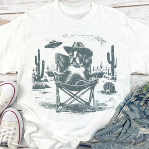 May include: White t-shirt featuring a graphic of a dog wearing a cowboy hat, relaxing in a camping chair in a desert scene with cacti and a UFO. The design is in shades of gray.