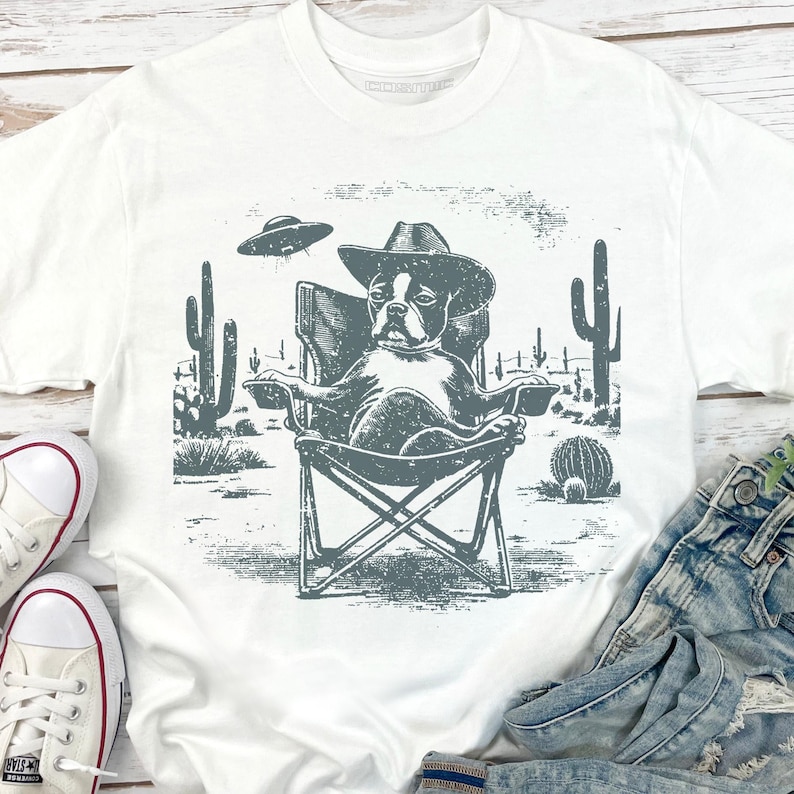 May include: White t-shirt featuring a graphic of a dog wearing a cowboy hat, relaxing in a camping chair in a desert scene with cacti and a UFO. The design is in shades of gray.