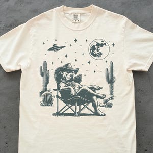 Pode incluir: Camiseta creme com um cachorro de chapéu de cowboy relaxando em uma cadeira dobrável sob uma lua cheia e um OVNI. O design inclui cactos e estrelas, criando um design caprichoso com tema deserto.