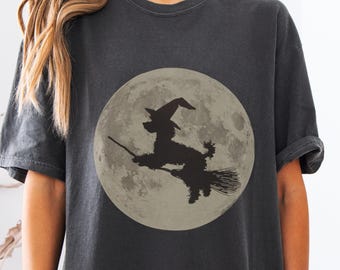 May include: Dark gray t-shirt featuring a full moon with a black silhouette of a witch riding a broom. The witch is wearing a pointed hat. The design is centered on the shirt, creating a Halloween theme.