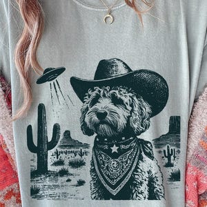 Könnte beinhalten: Hellgraues T-Shirt mit einem Hund, der einen Cowboyhut und ein Halstuch trägt, vor einer Wüstenlandschaft mit einem UFO. Der Hund hat eine Sternhalskette. Das Design ist in Schwarz- und Grüntönen gehalten.