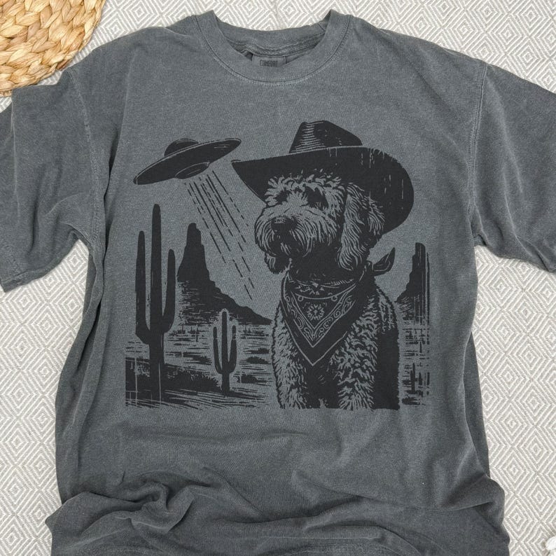 K&ouml;nnte beinhalten: Dunkelgraues T-Shirt mit einem schwarz-wei&szlig;en Grafikdesign. Das Design zeigt einen Hund mit Cowboyhut und Halstuch, mit einer UFO &uuml;ber einer W&uuml;stenlandschaft.