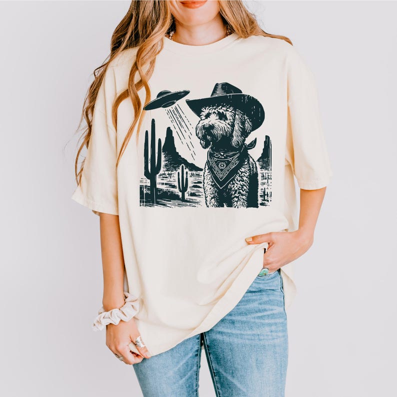 K&ouml;nnte beinhalten: Cremefarbenes T-Shirt mit einer schwarz-wei&szlig;en Grafik eines Hundes mit Cowboyhut und Halstuch, der von einem UFO entf&uuml;hrt wird. Der Hintergrund zeigt W&uuml;stenkakteen und Berge.