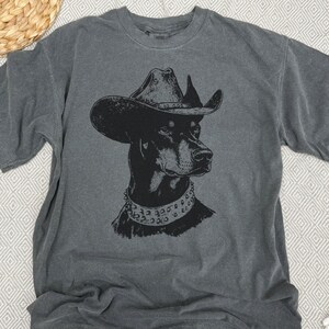 Pu&ograve; includere: T-shirt grigio antracite con grafica nera di un Dobermann Pinscher con cappello da cowboy e collare borchiato. Il ritratto del cane &egrave; centrato sulla maglietta, creando un design audace e accattivante.