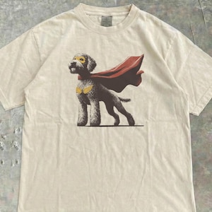 May include: Cream-colored t-shirt featuring a superhero dog graphic. The dog wears a yellow mask, gold wings, and a red cape. The graphic is centered on the shirt, which has short sleeves and a crew neck.