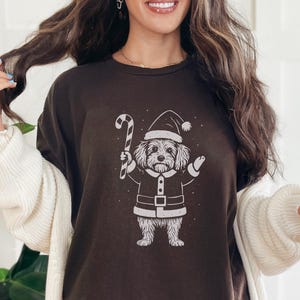 May include: A dark brown t-shirt with a white illustration of a dog in a Santa Claus outfit, holding a candy cane. The dog is wearing a Santa hat and suit. The shirt is a comfortable, casual style.