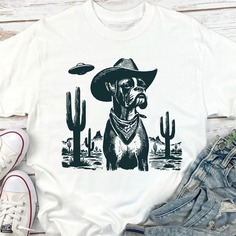 Puede incluir: Camiseta blanca con un gr&aacute;fico en blanco y negro de un perro Boxer con sombrero de vaquero y pa&ntilde;uelo, con un OVNI encima. El perro est&aacute; situado en un paisaje des&eacute;rtico con cactus y monta&ntilde;as.