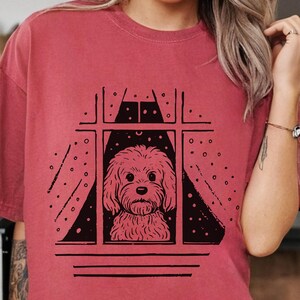 Op de afbeelding: Een roodroze t-shirt met een zwarte illustratie van een hond die uit een raam kijkt. De hond wordt omlijst door een raam met gordijnen en een sterrenhemel. Het t-shirt is effen van kleur.